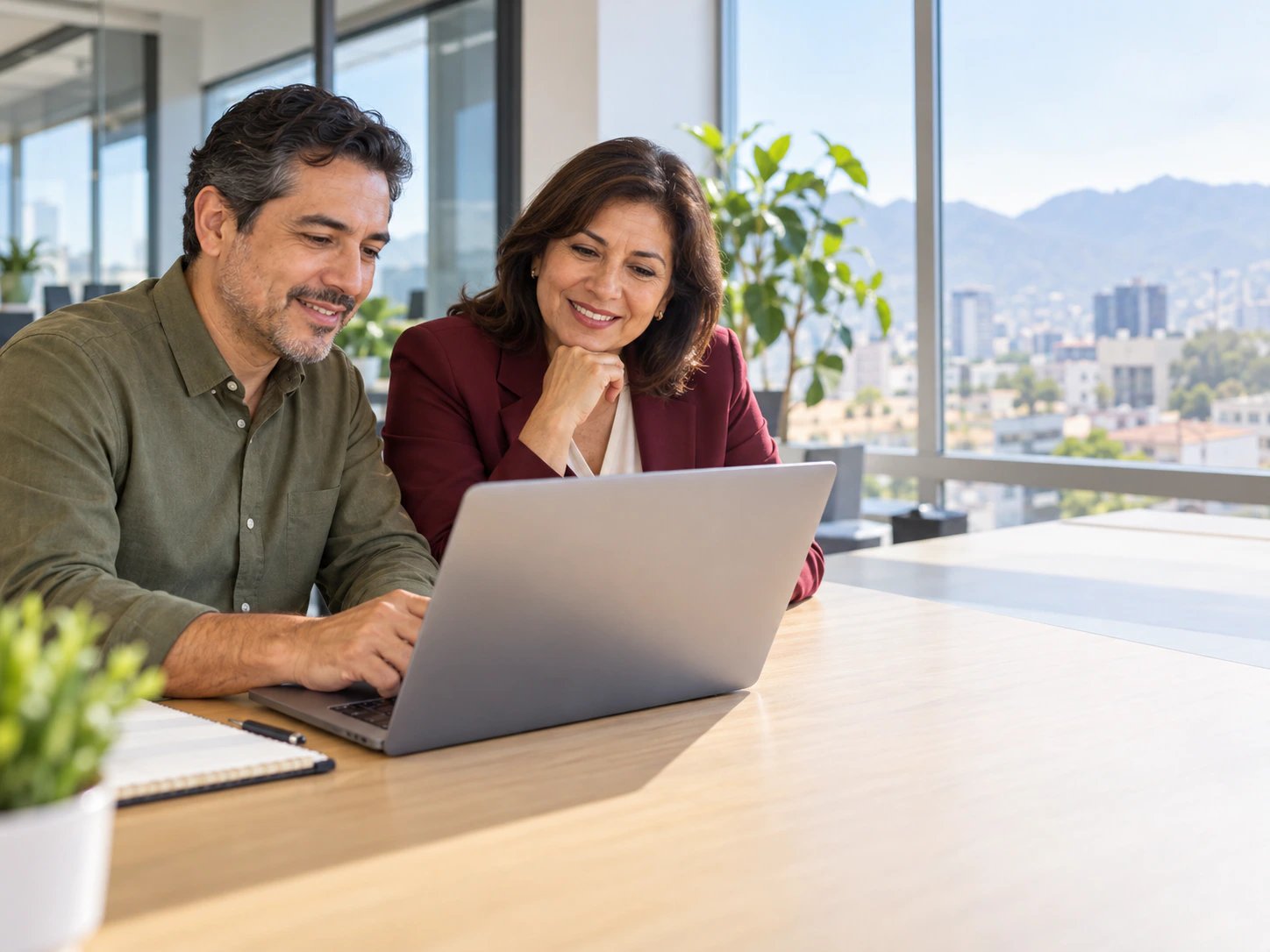 Dos profesionales revisando juntos un plan en una laptop, en una oficina con vista a la ciudad